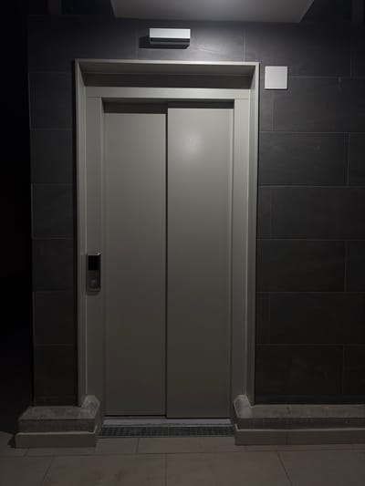 This image depicts a modern, minimalist bathroom or wet room area, possibly a shower or bathtub enclosure, in Cambrils Badia, Spain. The primary subject is the large, rectangular enclosure with a smooth, light grey surface. This surface appears to be a door or a panel, featuring two horizontal divisions. A small, dark electronic device with a screen and a circular icon is mounted on the lower horizontal section, suggesting it could be a control panel for the shower or lighting.

The enclosure is set against a backdrop of dark grey, large-format tiles that extend across the wall. To the left of the enclosure, a white, rectangular light fixture is mounted on the tiled wall. Above and slightly to the left of the enclosure, a small, square white plate is visible against the dark tiles, which might be a cover for an electrical outlet or a sensor.

The right side of the image reveals a concrete structure bordering the enclosure, with a slatted metal drain positioned next to it, further indicating a wet area. The overall lighting is dim, with a strong light source originating from the left, casting shadows and highlighting the textures of the materials. The scene is clean and functional, with a sense of understated luxury. No people are visible in the image. The time of day is not explicitly indicated but the artificial lighting suggests it could be evening or a time with low natural light.