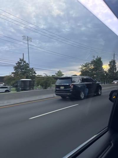 The image depicts a highway scene in Teaneck Township, New Jersey, captured from inside a moving vehicle. The time of day is characterized by an overcast sky with a patch of yellowish light near the right horizon, indicative of either early morning or late afternoon.
In the mid-ground, a dark-colored SUV, bearing a New Jersey license plate, travels in the same direction as the camera vehicle on a multi-lane highway. A reflection of a person's legs, clad in blue jeans, is visible on the side of the SUV, originating from the camera vehicle's interior. The highway lane is marked by a solid white line.
Beyond a concrete barrier to the left of the highway, two tall stadium-style light poles with multiple luminaires are prominent, suggesting an adjacent athletic facility. A small, dark, cylindrical utility structure stands behind the barrier on the left. Scattered trees with green foliage line the area behind the barrier. In the far left background, a white vehicle is partially visible, and a residential-style building is partially obscured by trees. Overhead utility lines crisscross the sky. In the far right background, another vehicle is faintly visible on the highway. The scene is consistent with the area surrounding Route 4 and the athletic fields of Teaneck High School or Votee Park in Teaneck.
