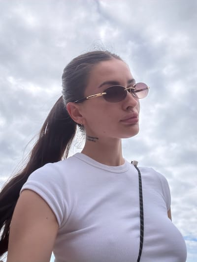 This is a close-up, low-angle, portrait-style photograph of a young woman wearing sunglasses and a white ribbed tank top. Her head is tilted back, and her gaze is directed upwards, suggesting a sense of contemplation or appreciation of the sky. The lighting suggests it is daytime, with diffused natural light indicating an overcast or cloudy sky, typical of a possibly humid or temperate climate.

The woman has dark hair pulled back into a ponytail, and she is wearing round, gold-rimmed sunglasses with tinted lenses. A thin black cord with white speckles is visible diagonally across her chest, possibly from a bag or clothing accessory. A small tattoo is visible on the back of her neck.

The background is dominated by a cloudy sky, with no discernible landscape or specific location cues beyond the provided context of Las Dehesas, Spain. The focus is tightly on the woman's face and upper body, emphasizing her expression and accessories. The overall mood is relaxed and candid. There is no visible text in the image.
