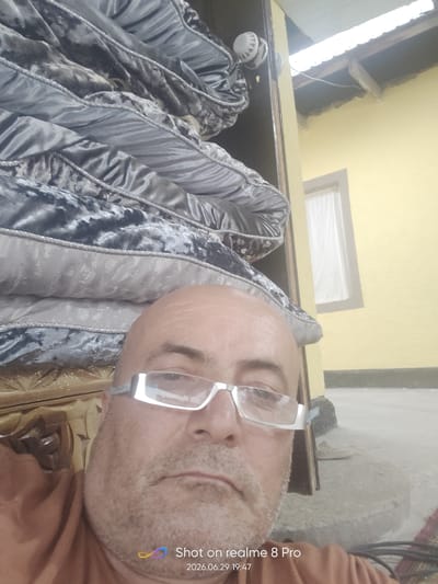 This is a close-up selfie taken outdoors, likely in Bokhtar, Tajikistan. The subject is a bald, middle-aged man wearing white-rimmed glasses and an orange-brown garment. He is positioned in the foreground, with a stack of large, dark gray and silver patterned pillows or mattresses behind him. To the right of the pillows is a doorframe and a yellow-painted wall, with a white-curtained window visible. The roofline of a building with exposed wooden beams is visible above the yellow wall. The ground appears to be concrete or dirt.
The lighting suggests it is daytime, possibly late afternoon given the slight shadows. The overall setting appears somewhat rustic or possibly under construction. The man's expression is neutral.
Visible text at the bottom right of the image reads "Shot on realme 8 Pro" and "2026.06.29 19:47".