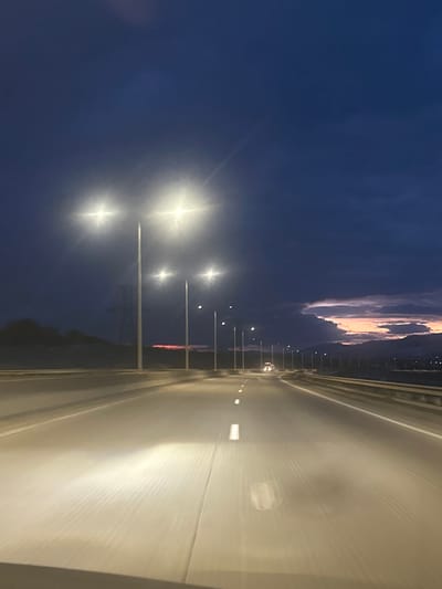 A nighttime view along a highway. Streetlights illuminate the left side, contrasting with the darkening blue sky above. Distant clouds show a hint of orange from a setting sun. The road surface is brightly lit, reflecting light into the camera, with occasional white lane markers. A vehicle is visible in the distance. The perspective suggests the image was taken from within a car.