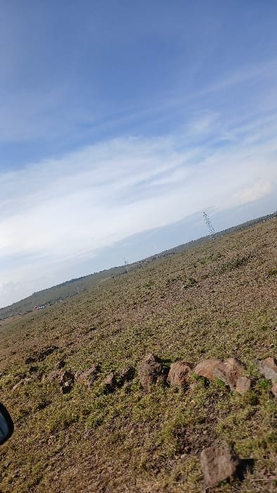 The image depicts an expansive, gently sloping open landscape under a partly cloudy sky, located within Kaputiei North ward, Kenya. The scene is captured with a noticeable upward tilt from left to right. The immediate foreground and midground consist of reddish-brown soil with sparse patches of low-lying green vegetation. A discernible line of irregularly shaped, dark brown rocks traverses horizontally across the lower-mid portion of the frame. Along the distant horizon, in the center-left, are faint indications of structures consistent with a small settlement or buildings. To the right of these, a series of vertically oriented power line pylons or communication towers extends into the distance. The sky, occupying approximately the upper two-thirds of the image, displays clear blue interspersed with scattered white cirrus and cumulus clouds. A partial view of a vehicle's side mirror is visible in the lower-left corner. No individuals, explicit actions, or direct interactions are observable.