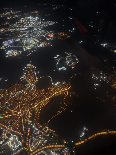 The image presents an aerial night view of the city of Tuzla, Turkey, captured from an aircraft. The scene is characterized by widespread artificial illumination against a dark background, revealing urban and coastal developments.

In the upper-center and central-right portions of the image, a prominent complex of bright white and bluish-white lights is visible, positively identified as Viaport Marina (Tuzla Marina) and its immediate surroundings. This area appears as a well-defined cluster of intense illumination, outlining structures, possibly buildings and entertainment zones, on a peninsula or coastline adjacent to a body of water. North-west of this complex, another extensive area of bright, structured illumination indicates a large commercial or industrial zone, consistent with Viaport Asia Outlet.

The lower-left and central parts of the image display a dense network of warm yellow and orange lights. These lights intricately trace roads and delineate residential areas, forming organic and grid-like patterns that represent the broader urban layout of Tuzla.

A dark expanse, consistent with the Marmara Sea, is visible, separating the intensely lit Viaport Marina complex from other landmasses.

A portion of a red airplane wing is present in the upper-right quadrant of the frame, confirming the viewpoint from an aircraft. No individuals or specific human actions are discernible due to the high altitude. The environment is dark, with ground illumination providing the sole visible light source below the aircraft.