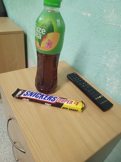 A bottle of Fuze Tea, a Snickers candy bar, and a black remote control are arranged on a light brown wooden bedside table. The bottle of Fuze Tea is green with a brown liquid inside. The Snickers bar is brown and yellow with the brand name "SNICKERS" in large blue letters. The remote control has many buttons and the brand name "Liberton" is visible. The background is a light green textured wall. The scene appears to be an indoor setting, possibly a bedroom or living room. There is no indication of the city of Khotsky, Ukraine.