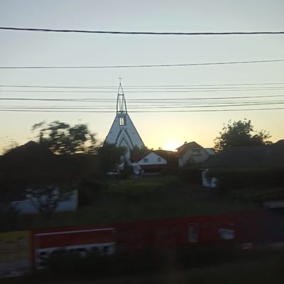 The image captures a wide-angle view of a village or town landscape in Nicolae Bălcescu, Romania, during what appears to be either sunrise or sunset. The sky is a pale, hazy gradient of light blue and yellow, with several horizontal power lines crisscrossing the upper portion of the frame.

Dominating the center of the image is the distinctive architecture of a church. It features a tall, pointed steeple with an open, lattice-like structure at its apex, topped by a cross. The main body of the church is triangular, painted white with dark, geometric patterns. Below the church, several residential buildings with red-tiled roofs are visible, interspersed with trees and foliage. Some of the trees are blurred, suggesting movement, possibly from the perspective of a moving vehicle.

The foreground is dark and out of focus, with a suggestion of a red and white horizontal structure, possibly a billboard or a building facade, running across the bottom of the image. The overall impression is that of a tranquil, rural or semi-rural setting bathed in soft, diffused light. No people are discernible in the image.