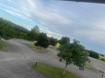 This is an eye-level, medium shot of a paved parking lot in Mainburg, Germany. The parking lot is made of gray cobblestones and is surrounded by green grass and trees. In the distance, there is a hill with more trees and some buildings. The sky is blue with some white clouds. The image is taken at an angle, giving a sense of depth and perspective. There are no people or animals visible in the image. A green tent-like structure is visible in the background.
