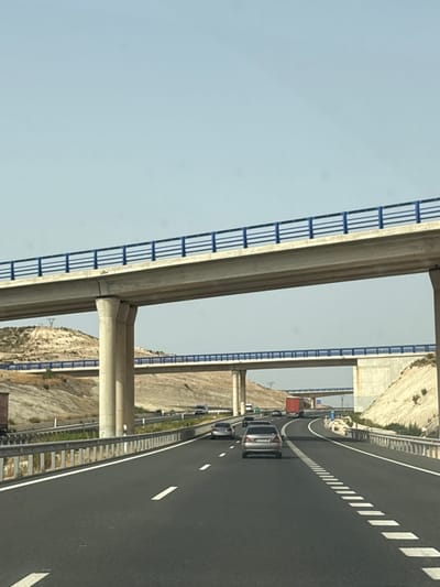 The image captures a highway overpass in Caudete, Spain, under a clear, pale blue sky. The foreground shows a black asphalt road with white dashed lines marking the lanes. Several cars, including a silver sedan and a dark-colored vehicle, are visible traveling on the road. In the midground, a multi-level highway structure is prominent. The upper level, supported by large concrete pillars, features a concrete roadway with a blue railing. Below it, another section of highway, also with a blue railing, runs horizontally. The landscape surrounding the highway is characterized by dry, eroded hills and vegetation. A red truck is visible on the lower highway. The scene is a typical depiction of a modern highway system, designed to facilitate vehicular traffic.