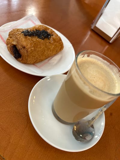 The image displays a close-up, elevated view of a meal setting on a reddish-brown, reflective tabletop within an establishment located in Vilanova I La Geltrú, Spain. In the foreground, a clear glass containing a light brown, frothy beverage, identified as coffee, rests on a white, round saucer. A small, silver-colored spoon with a textured handle is positioned on the saucer beside the glass. To the left and slightly behind the coffee, a golden-brown pastry, possibly a pain au chocolat, is presented on a white, oval plate. The pastry exhibits dark filling visible at one end and a sprinkling of dark particles on its top surface. A white paper doily with a red patterned border is situated beneath the pastry on the plate. In the upper right background, a chrome or clear acrylic napkin dispenser containing white paper napkins is partially visible. No individuals are present within the frame.