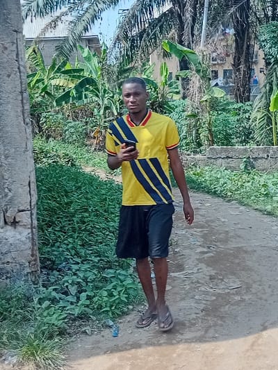 A single male individual is depicted standing on an unpaved dirt path in Ifite-Awka, Nigeria. He faces the viewer directly, holding a black smartphone in his left hand. The individual has dark skin and short, dark hair. He wears a yellow short-sleeved polo shirt with a red and black trimmed V-neck collar, featuring a pattern of diagonal black and blue stripes across the chest. He is also wearing black shorts and brown flip-flops.
The environment is an outdoor setting with dense green vegetation flanking the dirt path. Large-leafed plants, consistent with banana or plantain trees, are prominent, along with tall palm trees in the background. Partially visible behind the foliage are multi-story buildings. On the far left of the frame, a segment of a grey concrete or stone structure is present. A low concrete wall is visible in the mid-ground on the right. A piece of light blue plastic debris is on the path near the individual's left foot. The sky overhead is clear and blue.