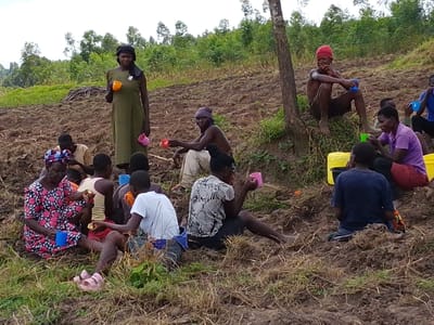 A group of people, primarily adults, rest on a brown, tilled hillside. Most are seated on the ground, each holding a colorful mug. A woman in a long green dress stands among them, also with a mug. The setting appears rural, with a wooded area visible in the background. A yellow water jug is present near a seated group, implying a break for refreshments. The atmosphere suggests a moment of communal rest during outdoor work.