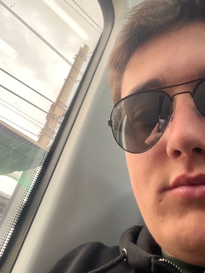 The image depicts a partial close-up of an individual, likely the photographer, positioned in the lower right half of the frame. The subject has light brown hair and is wearing dark aviator-style sunglasses, which reflect a hand holding a device (presumably the camera) and the interior of the vehicle. The subject is wearing a dark hooded sweatshirt over a green-collared garment, with their left cheek, nose, and lips partially visible. To the left, a vehicle window with a vertical dot matrix border reveals an exterior scene. Through the window, a large, light-colored stone building with a prominent classical facade, featuring multiple columns and architectural details, is visible against an overcast sky. Several parallel horizontal lines, possibly overhead wires, traverse the upper portion of the exterior view. A faint green object is visible in the lower left corner through the window. The building seen through the window is identified as the Església de Sant Cristòfol, located near the railway tracks in Premià de Mar, Spain. The individual is likely traveling on a train passing through Premià de Mar.