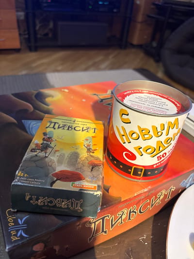 A cluttered table features several items. A cylindrical tin with Russian text, predominantly "С Новым Годом" (Happy New Year), sits atop a box of the board game "Dixit". Below the tin is a smaller, matching "Dixit" expansion pack box, labeled "Стиль жизни" (Style of Life). A larger, orange box, also seemingly for "Dixit", is positioned on the right. Part of a white plate is visible in the upper-right corner. A stand with a dark object on top is blurred in the background.
