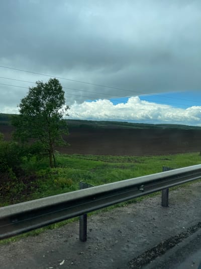 View from a moving vehicle, possibly on a highway, in Кокино, Russia. A metal guardrail is visible along the right side of the road, with a wet asphalt road surface. Beyond the guardrail is a strip of green vegetation, transitioning to a plowed field of dark soil. Further in the distance, a line of trees borders the field. In the sky, a mix of dark, overcast clouds and patches of bright white cumulus clouds are visible. A lone tree is partially visible in the foreground. Power lines stretch across the sky.