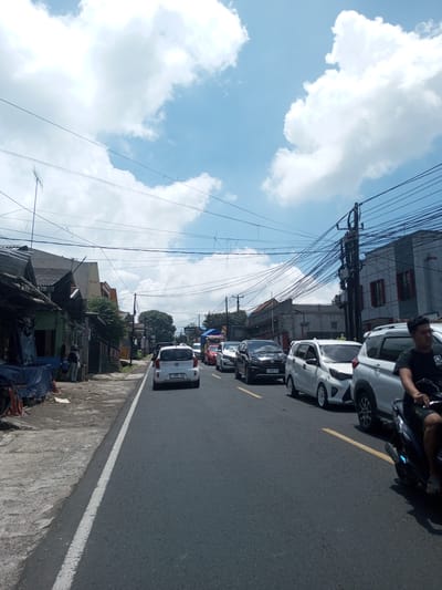 The image depicts a street scene in an urban area. Cars are lined up on the road, suggesting possible traffic congestion. A person on a motorbike is positioned on the right side of the frame, appearing to navigate the traffic. Buildings and overhead power lines are visible on both sides of the street, typical of a residential or commercial area. The weather appears sunny, with a blue sky and some clouds overhead. Based on the clues provided, the location might be in the vicinity of Kioumaknao 2, Laos, but this cannot be confirmed without explicit visual cues or landmarks.