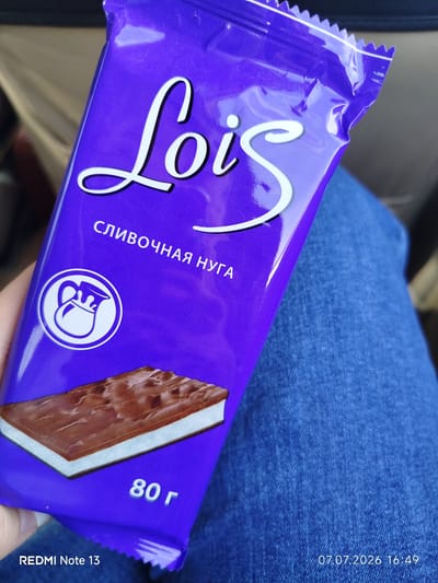 A close-up shot captures a purple chocolate bar wrapper with the white text "Lois" prominently displayed in a script font. Below the brand name, in smaller white text, is "СЛИВОЧНАЯ НУГА," which translates to "creamy nougat." To the left of this text, there's a circular white logo depicting a pitcher with milk pouring out. The bottom of the wrapper shows a graphic of the chocolate bar itself, revealing its nougat filling. The weight "80 г" is printed in white text on the lower right of the wrapper. The image also includes text overlays indicating "REDMI Note 13" and the date "07.07.2026 16:49." The chocolate bar is held by a person, and the background shows parts of blue denim clothing. The scene appears to be indoors, possibly in a car, given the muted, blurred background. There is no visible indication of the city of Nizhny Bestyakh, Russia.