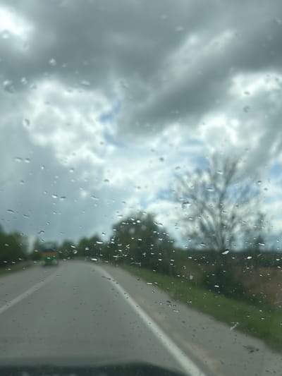 The image appears to be taken from inside a vehicle, looking through a rain-streaked window. Outside, a cloudy sky is visible along with blurry trees and greenery. Part of the vehicle's body is also in view, suggesting a close perspective. The location in Kiosevtsi, Bulgaria cannot be confirmed without additional visual clues in the image.