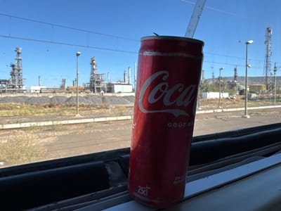 This image captures a close-up of a red 25cl Coca-Cola can with a white plastic straw, positioned on a dark window ledge, likely within a train or vehicle. The can features "Coca-Cola" and "GOUT ORIGINAL" in white lettering and shows surface moisture.

Beyond the window, which exhibits minor streaking, a clear blue sky is visible. The scene unfolds in an industrial area of Sidi Kacem, Morocco, specifically near the ONCF railway network, adjacent to what appears to be the former SAMIR oil refinery. Multiple railway tracks run horizontally across the midground. Adjacent to the tracks, an unpaved, dry earthen area is visible, containing sparse, light-colored vegetation and several conical piles of dark, coarse material, possibly gravel or industrial aggregate. A tall, slender grey light pole stands between the tracks and the distant industrial structures.

The background is dominated by a large industrial complex, characterized by numerous intricate metal towers, pipes, and storage tanks, indicative of an oil refinery or chemical plant. These structures are grey and metallic. Overhead, thin electrical wires traverse the sky. In the far distance on the right, faint, low-lying hills are discernible. The environment suggests a bright, sunny day. No human figures are present.
