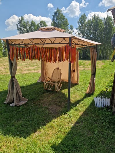 The image shows an outdoor scene on a sunny day, likely in Rikava, Latvia. The main subject is a tan-colored gazebo with a metal frame, set up on a grassy area. The gazebo is decorated with orange and yellow streamers hanging from its edges. Inside the gazebo, there is a wicker chair. The scene is viewed from a slightly elevated angle, looking towards the gazebo. The background features a line of trees and a bright blue sky with fluffy white clouds. A small section of a white picket fence is visible to the right of the gazebo. The grass appears well-maintained, with some areas showing shadows from the sun. The overall impression is one of a peaceful outdoor gathering space. There is no visible text in the image. The lighting suggests it is daytime, likely midday or early afternoon, given the strong shadows. The weather appears to be pleasant and sunny, ideal for outdoor activities.