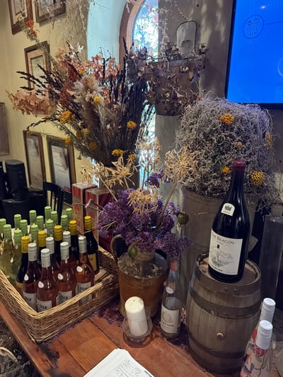 This image depicts a rustic and richly decorated interior scene, likely a wine tasting room, cellar, or shop, given the context of Drakenstein Nu, South Africa. The space is adorned with abundant dried foliage, including various grasses, leaves, and clusters of small yellow and purple flowers, creating a warm, autumnal aesthetic.

The central subjects are numerous wine bottles and wooden wine barrels. A prominent dark red wine bottle, labeled "THE BEACON SHIRAZ FAIRVIEW 2014," rests on its side atop a large wooden barrel. Below, a wicker basket is filled with multiple bottles, primarily labeled "ROAM," with light-colored labels and white caps, several also displaying "2025 AM." Another bottle, "FAIRVIEW QUARTZ," is also visible. The "Fairview" brand suggests a direct connection to the Fairview Wine and Cheese estate in the region.

In the background, a blue digital screen displays technical data, including "-4 kW Grid" and "2zero," juxtaposing modern technology with the rustic decor. The screen also shows text that appears to be "JAW GRIL." A rounded window with patterned or stained glass adds architectural detail. Other notable objects include framed pictures on the walls, a white candle in a holder, and a clipboard with a "VISITOR" sign and handwritten notes, further indicating a public or guest-oriented space. No people are visible, allowing the focus to remain on the curated display of wine and decorative elements.