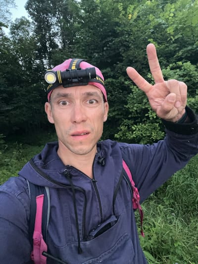 A man wearing a pink bandana with a headlamp is looking at the camera with a surprised expression. He is wearing a dark blue hooded jacket with a pink backpack strap visible. His right hand is raised, forming a peace sign. The background consists of green foliage and trees. The image appears to be a selfie taken outdoors, possibly in Il'metovo, Russia, given the context of the request.