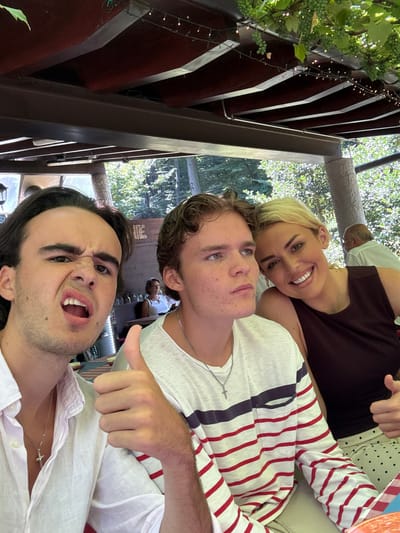 Three individuals are captured in a close-up selfie outdoors, likely at a restaurant or cafe. On the left, a young man with dark hair makes a humorous grimace, his mouth open and eyes squeezed shut. He is wearing a white linen shirt and a silver cross necklace. In the center, a young man with light brown hair looks slightly off-camera with a neutral expression, wearing a striped long-sleeved shirt and a silver chain. To the right, a woman with blonde hair is smiling broadly, her head resting on the shoulder of the young man in the center. She is wearing a dark sleeveless top and a polka-dot skirt. Behind them, other patrons are visible in the background, along with trees and a covered outdoor seating area adorned with string lights and grapevines. The table setting suggests a meal is in progress.