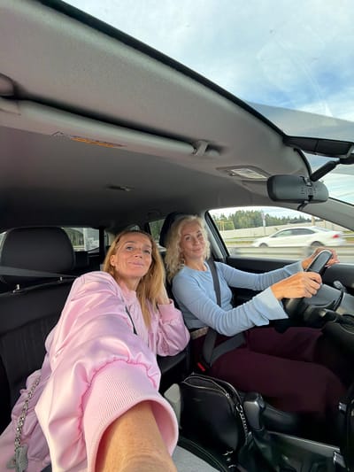 The image shows two women inside a car, seemingly in Russia. The woman in the driver's seat, wearing a light blue long-sleeved shirt, is holding the steering wheel and smiling. The second woman, in a pink jacket, is sitting in the passenger seat, also smiling at the camera. The interior of the car is visible, including the dashboard, steering wheel, and seats. Outside the car, trees and other vehicles can be seen, suggesting they are on a road. The weather appears to be overcast, with a bright sky visible through the windshield. The image is taken from an unusual perspective, possibly from the back seat or floor of the car, and is rotated.