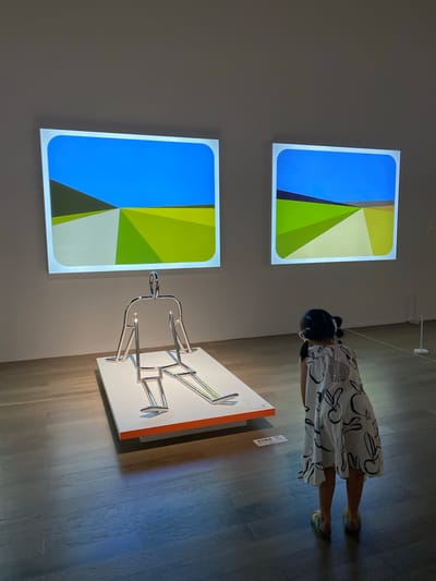 In the city of Chengdu, Jinniu District, a young girl stands with her back to the camera, facing two large, illuminated screens displaying abstract landscapes in shades of blue, green, and white. To her left, a chrome sculpture of a seated, humanoid figure is displayed on a white platform with an orange border. The girl, wearing a white dress with black rabbit prints and colorful sandals, is observing the art installation. The environment appears to be an art gallery with polished wooden flooring.