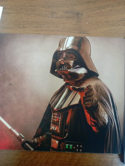 This is a close-up studio portrait of Darth Vader from Star Wars. He is depicted from the chest up, with his iconic helmet and cape. His right hand is extended forward, pointing directly at the viewer, with the lightsaber held in his left hand. The background is a smoky, reddish-grey gradient. The image is printed on a textured surface, likely canvas or paper, and is framed by a wooden surface at the top and bottom. There is no indication of the city of Ararat, Armenia within the image itself.
