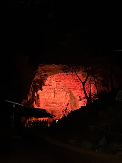 The image depicts a dramatic scene within a cave, likely a tourist attraction, illuminated by a vibrant red light. The light casts a warm glow on the rough, textured rock formations of the cave walls and ceiling, highlighting their stratification and uneven surfaces. In the foreground, a dark, silhouetted cluster of people is visible, suggesting a group of visitors observing the illuminated cave. The people are indistinct and appear to be gathered together, possibly listening to a guide or simply taking in the spectacle. To the left of the group, a large, dark object, possibly an umbrella or awning, is partially visible, extending into the frame and casting its own shadow. A thin, dark tree trunk with branches is silhouetted against the red light on the right side of the image, adding a natural element to the subterranean landscape. The overall impression is one of awe and wonder, with the intense red lighting creating a theatrical and somewhat mysterious atmosphere within the natural confines of the cave. The location within the city of Gollagutta, India, is not directly evident from the visual information provided in the image itself, and would require external contextual knowledge.