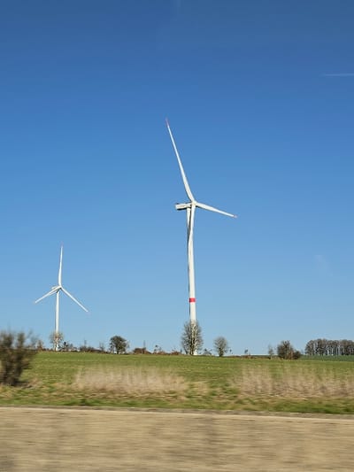 An exterior scene in Amberloup, Belgium, depicts two white industrial wind turbines against a clear blue sky during daylight. The foreground presents a horizontal band of blurred, light brown and tan ground, consistent with motion blur. Above this, a green grassy field stretches across the midground. The larger, closer wind turbine on the right features a white tower with a single red horizontal band and three blades, one pointed upwards with a red tip, and two horizontally. A smaller, more distant wind turbine on the left also has a white tower and three blades, one pointed upwards with a red tip. Sparse, bare deciduous trees are visible on the horizon line between and around the turbines, with a denser treeline present on the far right. No individuals or discernible actions are present.