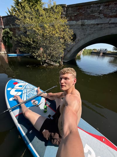 A medium shot captures a shirtless man on a paddleboard in the River Stour, taken in Babergh, United Kingdom. The man, positioned centrally, is seated on the board with a paddle in his hands. A bottle and sunglasses rest on the board near him. He is tan, short blond hair. He is turned towards the camera, looking directly at the viewer. The paddleboard is blue and white, with a distinct design on its front. The river appears calm, with dark, still water. Behind the man, a stone bridge arches over the river, framing a view of more water and some vegetation beyond. The bridge's architecture includes brickwork and some greenery growing on its surface. A tree with light green leaves is visible to the left of the bridge. The sky above is clear and blue.