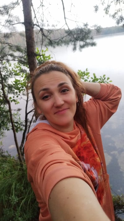 A selfie taken outdoors shows a woman with auburn hair and a light complexion. She is wearing an orange hooded sweatshirt with a floral print on the inside of the hood. Her right arm is raised, with her hand touching the back of her head. She is smiling slightly at the camera. The background consists of greenery, trees, and a body of water, likely a lake or river, under an overcast sky. The image does not provide enough information to determine the specific location within Tarasovskoe, Russia.