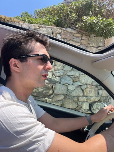A male individual is depicted in the driver's seat of a vehicle with an open sunroof, located in an outdoor setting in Agios Ioannis Diakoftis, Greece. The subject, positioned in profile facing right, has dark, curly hair and wears dark sunglasses featuring a white star pattern on the frames. He is dressed in a light-colored, ribbed polo shirt. Red marks are visible on the left side of his neck. His left hand, adorned with a watch featuring a grey strap and gold-colored bezel, rests on a light-colored steering wheel. The background consists of a stone wall composed of irregular light-colored stones, topped by green vegetation, under a clear blue sky. The scene is illuminated by natural light.