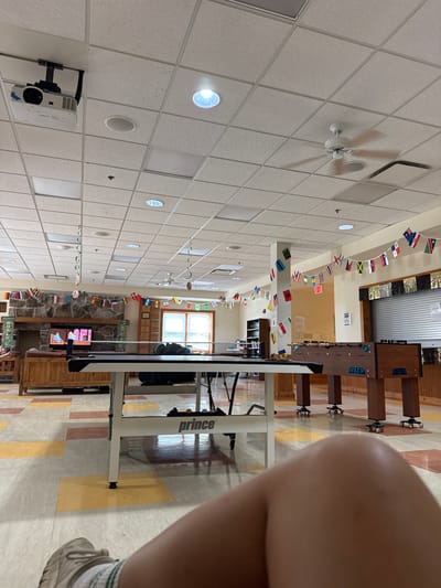 The image shows a room with a tiled ceiling, featuring circular lights and a projector mounted on the ceiling. A string of small flags from various countries is draped across the room. In the background, a foosball table is visible, and a ping pong table is also present. A stone fireplace with a television screen above it is also in view. The floor has a patterned design with rectangular blocks of color. A person's leg and foot, wearing athletic shoes, are in the foreground, obscuring part of the scene. The lighting suggests it is daytime, and the setting appears to be an indoor recreational area or common space, possibly a school or community center in Washington Township, USA. There is no discernible weather or emotional context. Visible text includes "prince" on the ping pong table and some unreadable text on the foosball table and projector.