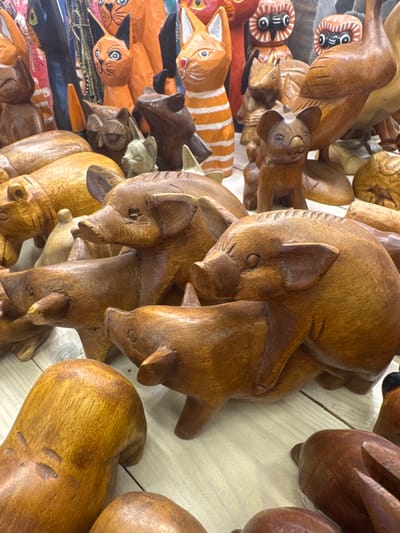 The image displays a close-up, overhead view of numerous carved wooden animals, predominantly cats, arranged closely together. The scene appears to be a display of handicrafts, possibly in a market or shop. The carvings vary in size and detail, with some featuring painted accents like stripes or facial features, while others are left in natural wood tones. Some carvings are stacked on top of others, creating a layered effect. The lighting suggests an indoor setting or a shaded outdoor area. The overall impression is one of artisanal craftsmanship and a collection of decorative items.