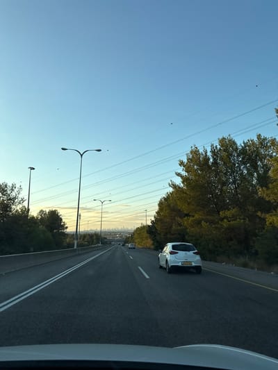 The image depicts a multi-lane highway scene, observed from inside a vehicle, presumably in the Kerem Ben Shemen area of Israel, looking west towards a distant city skyline. The time of day appears to be late afternoon or early evening, characterized by a clear light blue sky transitioning to a yellowish hue at the horizon.

In the foreground, the top of a vehicle's dashboard or hood is visible. The midground features a paved highway with two visible lanes of travel marked by double white lines, and a solid yellow line delineating the shoulder on the right. A white hatchback vehicle, identified as a Mazda 3 based on its design, is centrally positioned in the right lane, moving away from the viewer. Its rear license plate, partially obscured, displays "643:21-701". Several other vehicles are visible further down the highway, also traveling in the same direction. Tall streetlights with bifurcated arms are positioned along the highway, primarily on the left side. Dense vegetation, including coniferous trees, borders both sides of the road, forming a significant forest line on the right.

In the background, multiple parallel power lines equipped with spherical orange/red marker balls traverse the sky. Beneath these power lines, a distinct urban skyline, consistent with Tel Aviv, is visible on the distant horizon. No people are discernible in the scene.