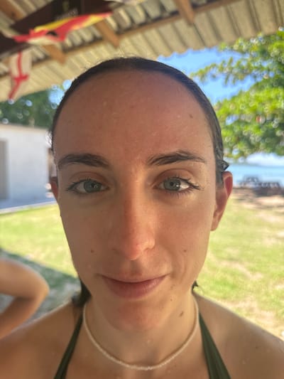 A female subject is centered in a close-up, head-on portrait, with fair skin, dark eyebrows, and light blue-green eyes. Her dark hair is pulled back and appears wet, along with visible water droplets on her face and shoulders. She wears a dark green swimsuit top and a thin white beaded necklace, displaying a neutral to slightly smiling expression. The scene is set in Rio Bueno, Jamaica. In the upper background, under a wooden beam roof, a red, yellow, and black banner (partially identifying as Guinness) and a white flag with a red cross are visible. The background, out of focus, features a white building to the left, green foliage and trees to the right, and a body of water with boats in the far distance, beneath a clear blue sky. Green grass occupies the mid-ground. The blurred arm of another individual is partially visible in the lower-left.