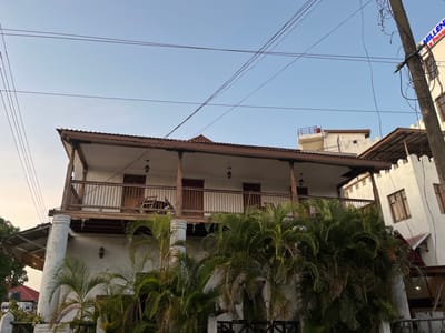 The media file presents an exterior view of a two-story building, likely a hotel, under a clear, light blue sky, suggesting either early morning or late afternoon. The main building features white stucco walls and a prominent wooden balcony on the second floor, complete with a detailed wooden railing and support columns. The balcony roof is dark brown with ornate wooden eaves. On the balcony, a wooden chair is visible. Below the balcony, large, rounded white columns support the structure, and a light fixture adorns the ground floor wall.
In front of the building, lush green palm trees partially obscure the lower facade. To the right of the main building, an adjacent white structure with battlements along its roofline is visible, sharing a similar architectural style. Farther right, a utility pole with numerous black electrical wires cuts across the frame.
Visible text on a sign partially obscured by foliage at the bottom left reads "MILLENNIUM LA RENAISSAN HOTEL," identifying the establishment. A portion of the same sign, "MILLENNI" and "E RENAI", is also visible on an upper building to the far right. The overall scene, with its tropical foliage and distinct architecture, aligns with the provided location context of Bagamoyo, Tanzania. No people or specific activities are depicted.