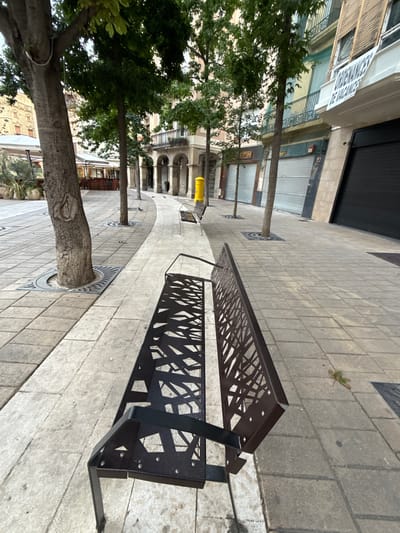 This is a wide-angle, ground-level shot of a pedestrian walkway in Igualada, Spain, with a modern black metal bench in the foreground. The bench has a geometric, cutout design on its backrest and seat. The walkway is paved with light-colored rectangular tiles.
To the left, large trees with rough bark are planted in circular grates with a wave pattern. Beyond the trees, there are white umbrellas shading outdoor seating areas, possibly for cafes or restaurants, with wooden chairs and tables visible. Further in the background, the facade of a building with arches and balconies can be seen.
To the right, the walkway continues, and another black metal bench is visible in the mid-ground, similar in design to the one in the foreground. A bright yellow cylindrical object, possibly a public bin or bollard, stands near this bench. The buildings lining the right side of the walkway have a colorful facade with balconies and shuttered windows. A white banner with text, including "ORDENANCES DE VACANCES," is hanging from a balcony. The ground level of these buildings appears to be commercial spaces with closed metallic roll-up doors.
The lighting suggests it might be late afternoon or early morning, with shadows cast on the ground from the trees and benches. A few pigeons are scattered on the walkway. The overall scene depicts a clean and well-maintained urban public space.