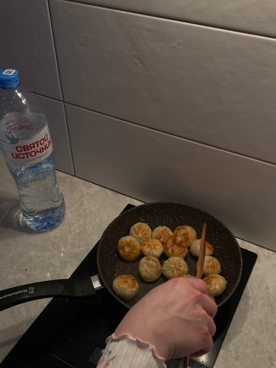 The image shows a pan with dumplings being cooked on a stovetop. A hand is using chopsticks to move the dumplings. A bottle of "Святой Источник" water is on the counter near the stovetop. The location is Зубово, Russia.