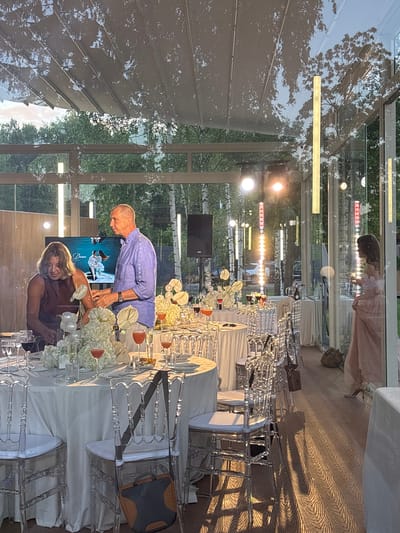 The image depicts an outdoor event space with a glass enclosure. In the foreground, several round tables are set for a meal, adorned with white tablecloths, floral arrangements, and glassware. Transparent chairs surround the tables. A man and a woman are standing by one of the tables; the man is wearing a blue shirt, and the woman is wearing a dark-colored dress. A large screen behind them displays an image of a couple. The background reveals trees and what appears to be other parts of the venue, including lighting fixtures and more tables. The scene is illuminated by natural light and artificial lighting, creating reflections on the glass surfaces. The floor is made of wood. The setting suggests a celebration or a formal gathering.