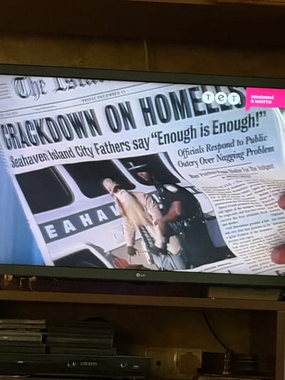 The image shows a television screen displaying a news article from a newspaper. The headline reads "CRACKDOWN ON HOMELESS". There is a smaller headline that says "Seahaven Island City Fathers say 'Enough is Enough!'". The article is printed in black ink on what appears to be a white or off-white newspaper. Below the headlines, there is an image of a police officer in uniform, appearing to interact with a person. The person is partially obscured and appears to be wearing civilian clothing. The background of the newspaper article features a graphic of a bus or train with the letters "E A H A V" visible. In the top right corner of the screen, there are three circular icons with the letters "т е т" inside them, and a pink speech bubble with Ukrainian text "закохані в життя" which translates to "in love with life". The television is placed on a shelf, and a stack of DVDs or Blu-rays is visible below it. The overall setting appears to be a domestic environment, likely a living room. No specific location in Osolynka, Ukraine is visible or suggested by the image content.