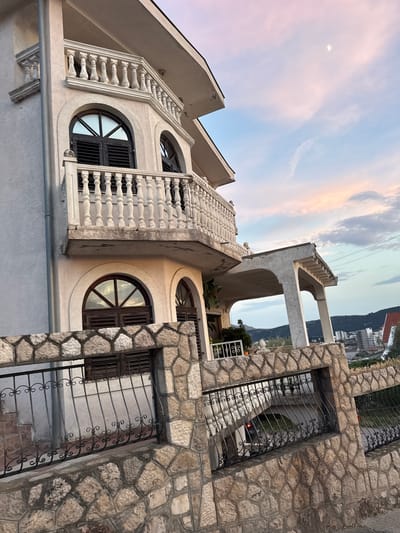 The image shows a multi-story white building with decorative balconies and arched windows, set against a sky with soft pink and blue hues, suggesting either dawn or dusk. The building's architecture features ornate balustrades and stone accents. In the foreground, a stone wall with an ornate black metal fence borders the property. Beyond the building, a city skyline with various buildings and hills is visible. The overall scene is a residential area in the city of Burtaiši, Montenegro. There are no people or discernible actions captured in the image.