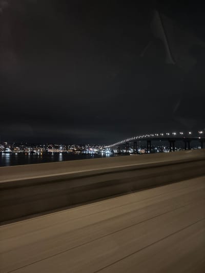 The image depicts a nighttime cityscape view, potentially observed from an elevated position within a building or from a window. The location appears to be Coronado, United States of America, as identified by the presence of the San Diego-Coronado Bridge illuminated with a continuous string of lights, stretching horizontally across the middle portion of the image. Various city lights can be seen in the foreground. A section of window or perhaps a closed curtain occupies the right side of the frame, adding depth and serving as a visual barrier or boundary to the scene. The scene shows a body of water with reflected light. The sky is dark and featureless.