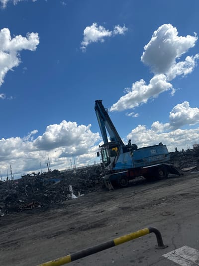 The image depicts a blue Terex material handler crane positioned at a scrap yard in Dzerzhinsky, Russia. The crane, featuring a prominent boom and a driver's cabin, is partially visible against a backdrop of a vast pile of scrap metal. The scene is illuminated by bright daylight, suggesting it is daytime. The weather appears clear with a vivid blue sky dotted with fluffy white cumulus clouds. In the foreground, a concrete surface is visible, marked with a yellow and black striped pole, likely a barrier or marker. The overall setting is industrial, characterized by the presence of heavy machinery and accumulated scrap materials. The image is oriented vertically, giving a sense of height and scale to the crane and the surrounding environment. No people are clearly visible in the image.