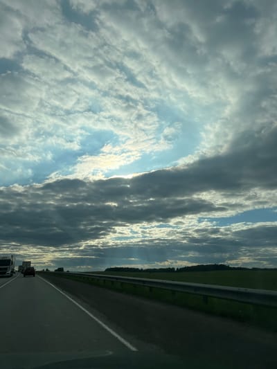 The image shows a highway scene with a dark, cloudy sky and some patches of blue. The sun is partially obscured by clouds, casting rays of light downwards. On the highway, there are several vehicles, including a white truck on the left, a red car in the middle distance, and other trucks further ahead. A guardrail runs along the right side of the highway, separating it from a green field. The landscape in the background consists of fields and trees, suggesting a rural or semi-rural location. The weather appears to be overcast with some dramatic lighting from the sun breaking through. The location is identified as Сухая Балка, Russia.