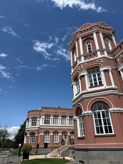 The image depicts a large, ornate building with terracotta-colored walls and white trim, possibly part of a palace or historical estate, located in Mukhrani, Georgia. The building features tall, arched windows and a prominent turret with crenellations. The sky is a vibrant blue, dotted with sparse white clouds. In the foreground, on a paved pathway, are a few white tables and a dark speaker on a stand. A manicured lawn borders the pathway, with some trees and greenery visible. To the left, a section of another similar building is visible, and in the distance, a few individuals can be seen walking. The architectural style suggests a classical European influence.