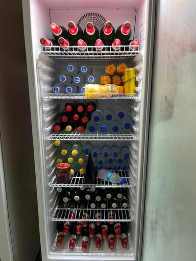 The image shows an open commercial refrigerator filled with various beverages. The refrigerator is brightly lit, suggesting it is daytime. The scene is an indoor setting, likely a store or restaurant, given the organized display of drinks.
In the left section of the refrigerator, there are several dark green bottles with red caps and labels that read "DRAFT BEER". These bottles are arranged vertically. Next to them, in a separate compartment, are yellow bottles of "Sodawater" and orange bottles of "Pridina" juice.
The middle section of the refrigerator contains numerous clear plastic bottles filled with water, each with a blue cap. Some bottles have labels with "Pocari Sweat" written on them. There are also rows of dark colored bottles with red and yellow caps, likely soft drinks, and some clear bottles with yellow caps.
The right section of the refrigerator is primarily filled with more clear plastic water bottles with blue caps. Towards the back of this section, there are rows of dark bottles with red and white labels, which appear to be wine bottles. One bottle in the middle of the right section has a red lid and a darker colored body.
The overall impression is one of a well-stocked beverage cooler. There are no people visible in the image, and the focus is entirely on the arrangement and variety of drinks. The metal racks and white interior of the refrigerator are clearly visible. The lighting is even, and there are no strong shadows, indicating a controlled indoor environment.