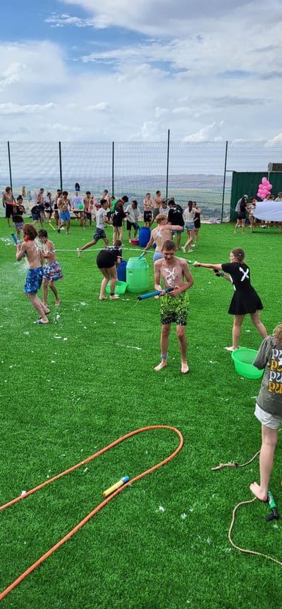 The image depicts a lively outdoor water fight or party taking place on a bright, cloudy day. The scene is set on an artificial turf field, likely a sports ground or recreational area. A metal fence encloses the area, with a blurred landscape of hills and a few structures visible in the background.
Numerous individuals, primarily young people and teenagers, are engaged in water-based activities. Many are holding water guns and appear to be spraying water at each other. Some individuals have white markings or paint on their bare chests, suggesting a game or a pre-event activity. Various water containers, including large green barrels and smaller buckets, are scattered across the field, some filled with water.
In the foreground, an orange hose lies coiled on the turf, and a yellow and blue water gun attachment is visible near its end. A rope is also present, suggesting other potential activities. In the far right of the image, a person wearing a grey t-shirt with a patterned print and white shorts is partially visible, facing away from the camera.
The overall atmosphere appears to be one of fun and energetic play. The location is identified as Сырымбет, Kazakhstan.