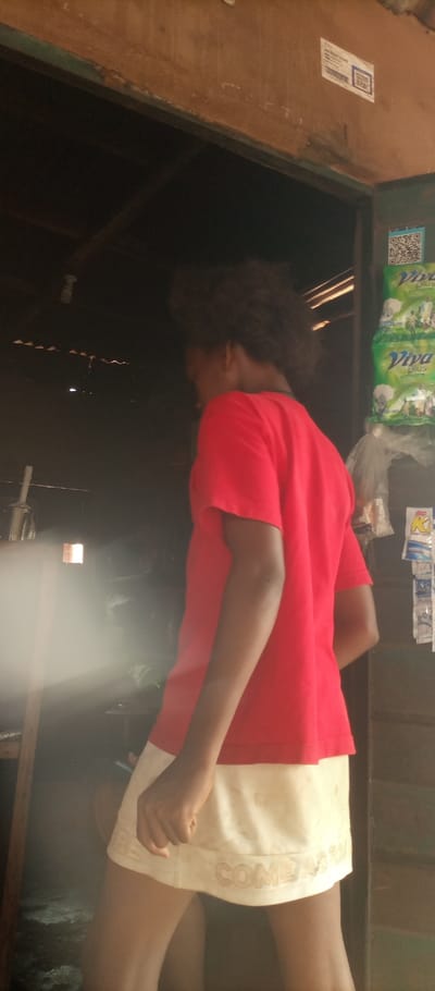 A person with dark, curly hair, wearing a red short-sleeved t-shirt and an off-white skirt or shorts (bearing text "COMEAS" on the hem), stands in a doorway in Umuoji, Nigeria. The individual is positioned with their back partially to the camera, looking into a darker interior. Their left arm is visible, hanging by their side with a partially clenched fist. To the right of the person, a wooden door frame or wall displays multiple green plastic pouches (labeled "Viva Plus") and clear plastic bags containing various sachets. Two white stickers with QR codes are affixed to the wooden structure. The interior to the person's left is dimly lit, featuring a hanging light fixture and a significant volume of white mist or steam. Indistinguishable objects are visible behind the mist. An overhead wooden beam forms the top of the frame.